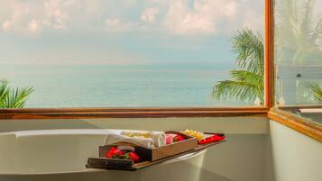 Eine Badewanne mit Tablett, auf dem Pflegeprodukte und ein Handtuch liegen, mit Blick auf ein Gewässer und Palmen.