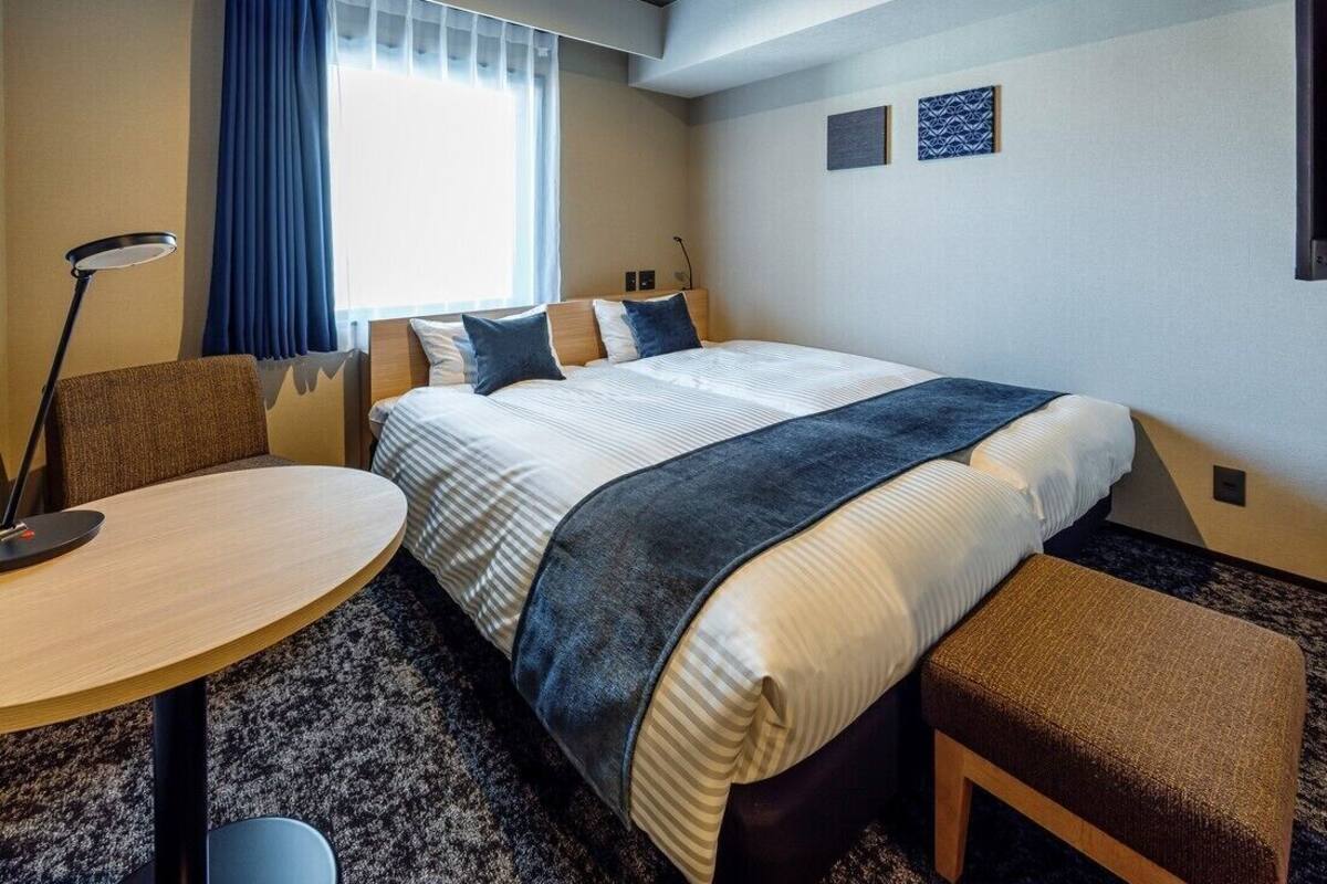 A hotel room with a bed, a desk, a chair, a small table, a bench, a lamp, and a window with curtains.