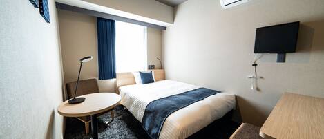 A hotel room with a bed, a desk, a chair, a desk lamp, a TV, and an air conditioning unit.