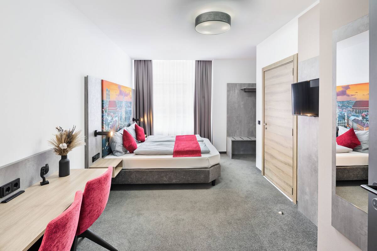 A modern hotel room with a bed, a desk, a chair, a TV, and artwork on the walls.
