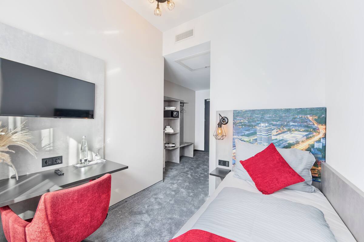 A modern hotel room with a flat-screen TV, a desk with a chair, a bed with red and white bedding, and a cityscape painting on the wall.