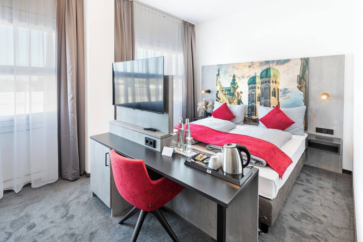 A hotel room with a bed, a desk, a TV, and a painting of a building on the wall.