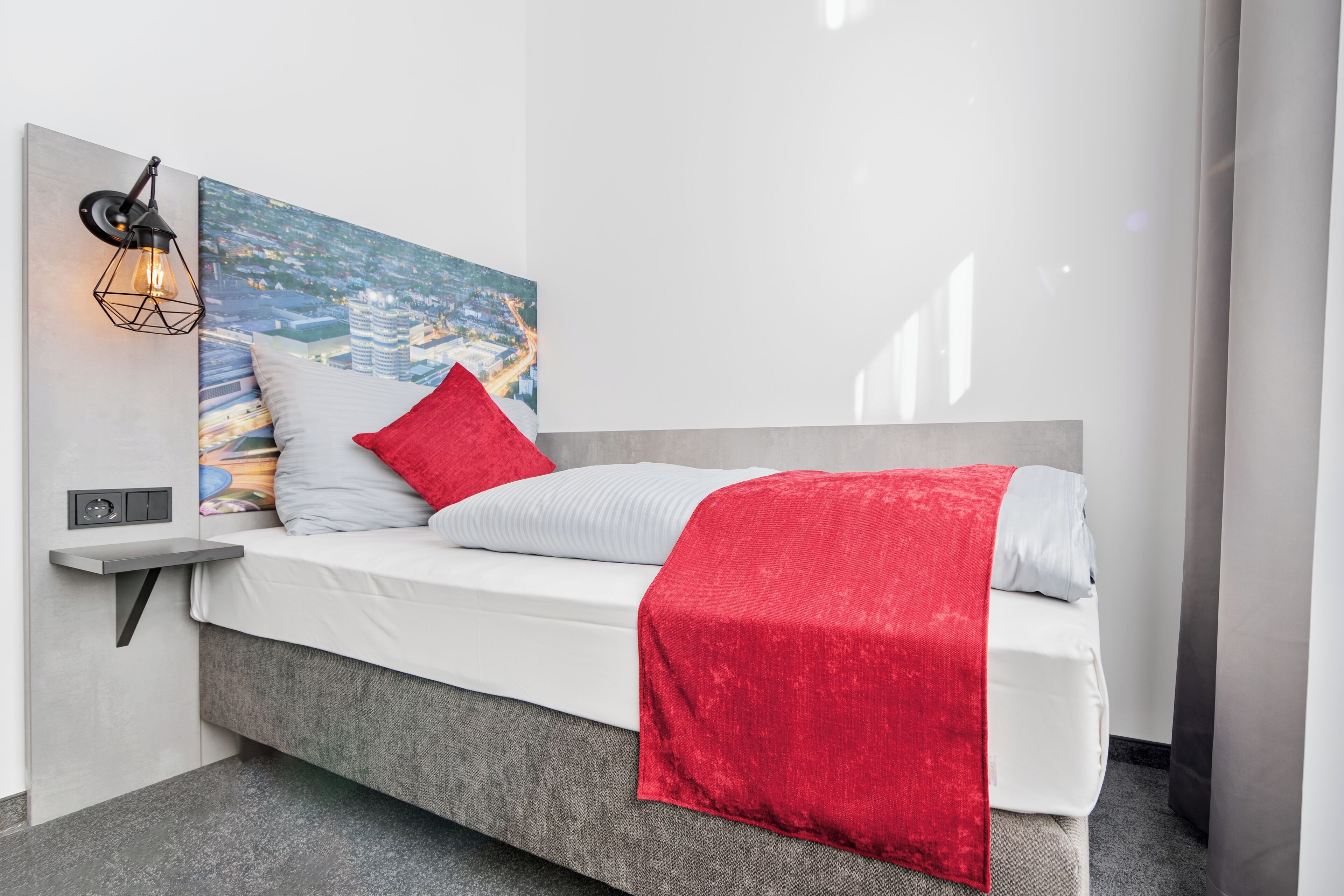 A modern bedroom with a bed, a red throw blanket, a cityscape painting, and a wall-mounted light fixture.