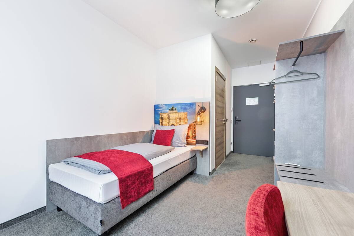 A single bed with a red blanket, a painting on the wall, and a wardrobe with a hanger.