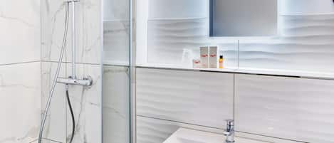 Un baño moderno con ducha de vidrio, un lavamanos blanco con espejo arriba y una pared de azulejos con un diseño que imita el mármol.