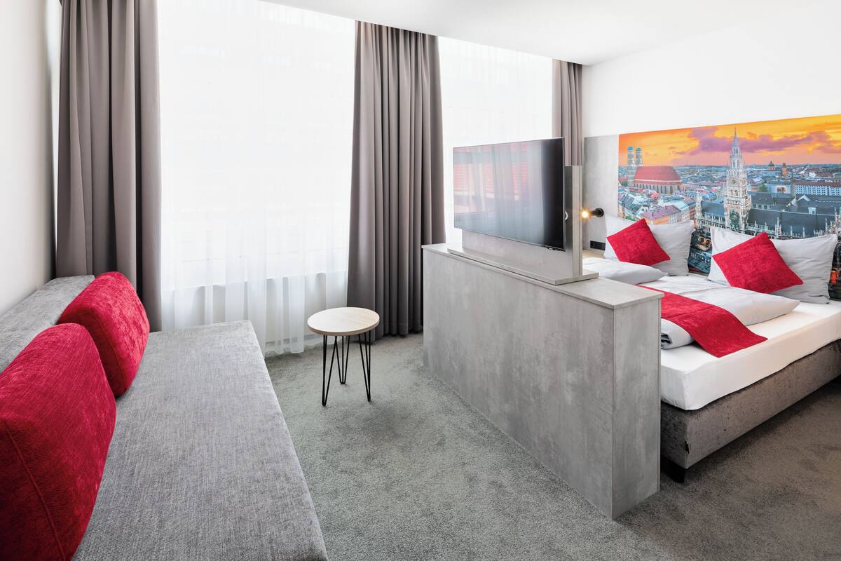 A modern hotel room with a grey sofa, a small round table, a bed with red and white bedding, and a cityscape painting on the wall.