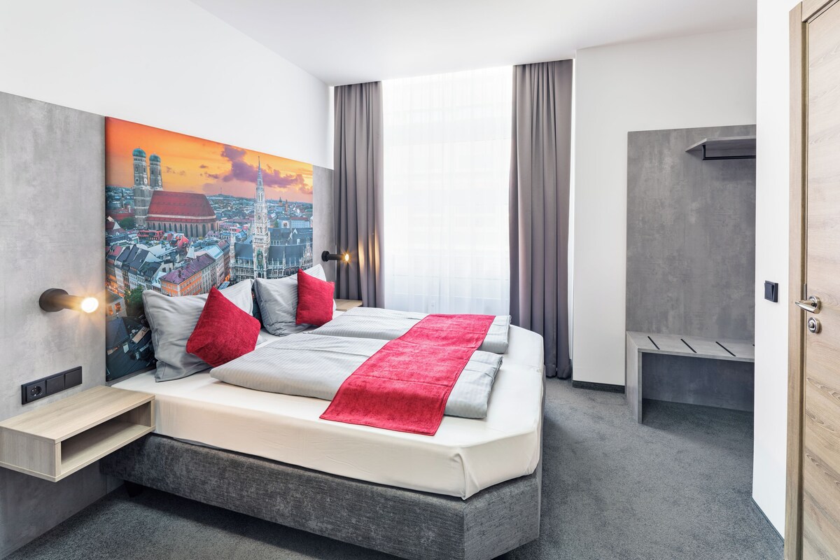 Quarto de hotel moderno com uma cama grande, uma pintura de paisagem urbana na parede, uma mesinha de cabeceira e vista para a cidade.