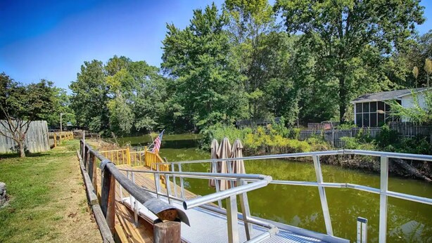 Une terrasse en bois, agrémentée de rampes métalliques, surplombe une étendue d’eau calme, le tout étant entouré d’une végétation luxuriante et d’arbres.