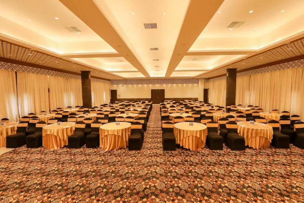 Een ruime congreszaal met rijen tafels en stoelen, een kamerbreed tapijt en een hoog plafond met inbouwverlichting.
