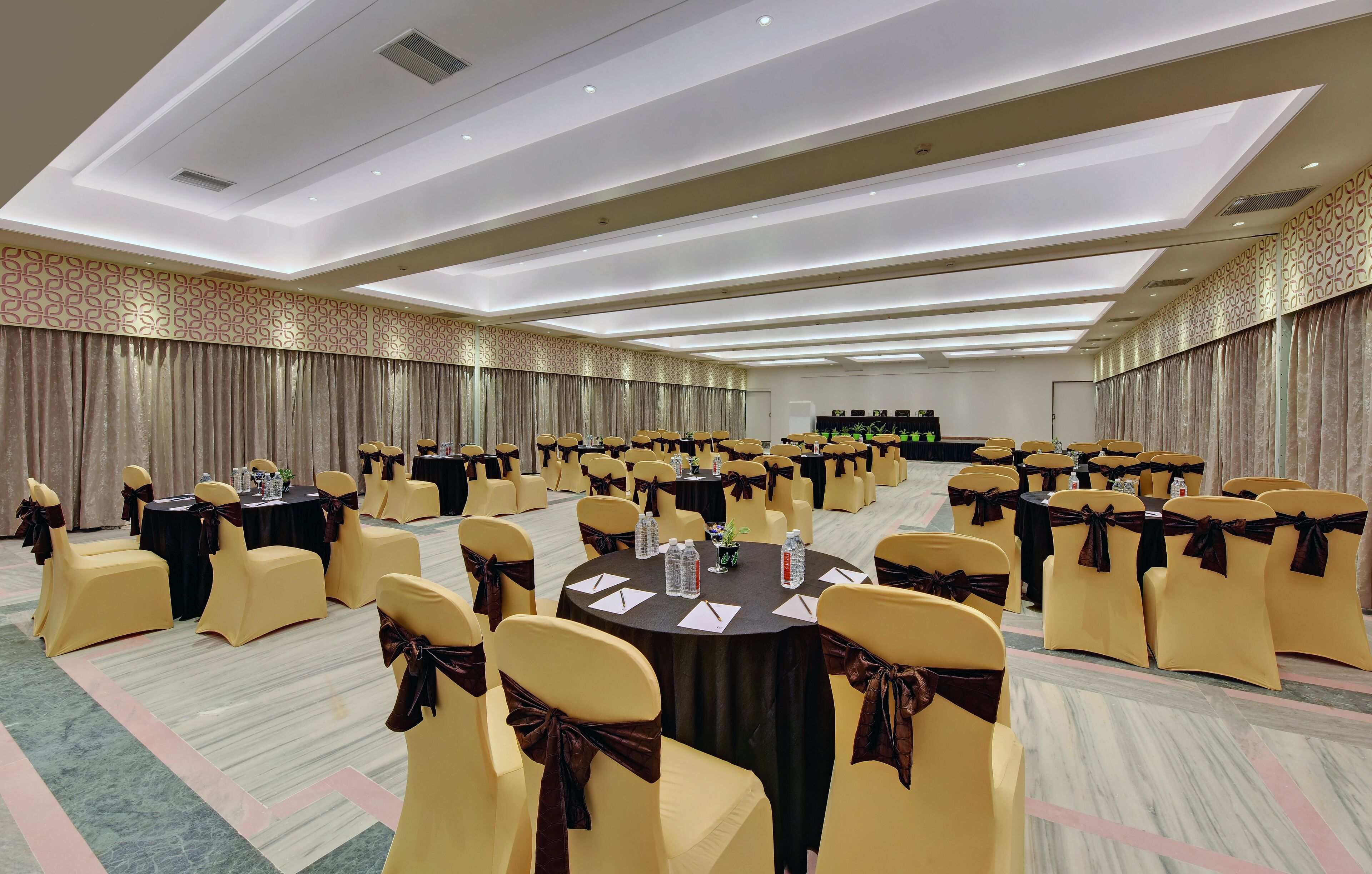 A large conference room with round tables, yellow chairs with brown sashes, and a patterned wall.