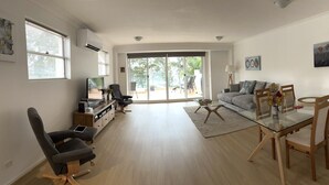Un salón espacioso con un gran ventanal corredera que da a un balcón, un televisor de pantalla plana, un sofá, una mesa de comedor con sillas y un mueble de madera.
