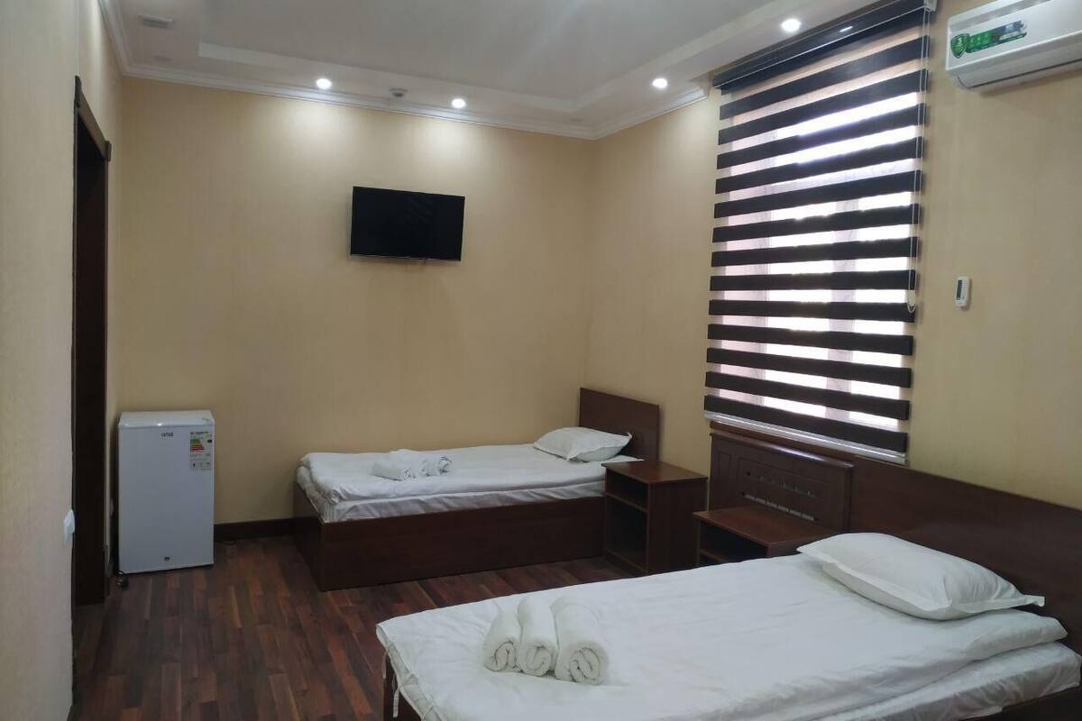 A hotel room with two single beds, a small refrigerator, a flat-screen TV mounted on the wall, and a window with blinds.