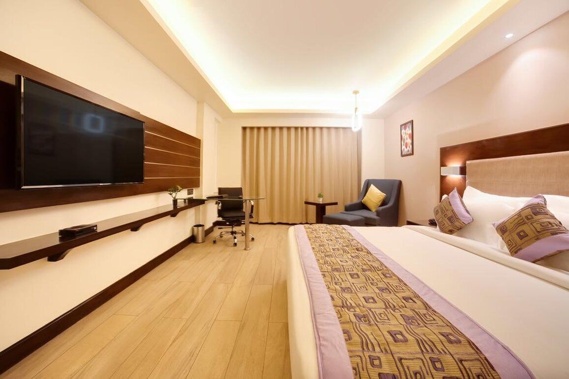 A hotel room with a large flat-screen TV mounted on the wall, a bed with patterned bedding, a desk with a chair, and a small table with a lamp.