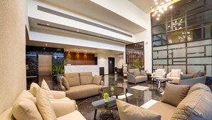 A spacious lobby with a modern design, featuring a large seating area with sofas and armchairs, a central coffee table, and a reception desk.