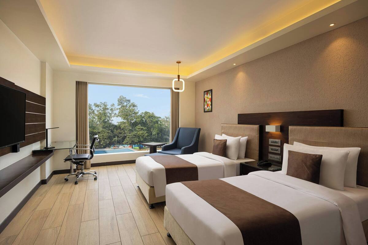 A hotel room with two beds, a desk, a chair, a TV, and a view of the outdoors.