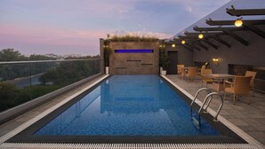 A rooftop pool with clear glass walls, a blue-lit water feature, and outdoor seating.