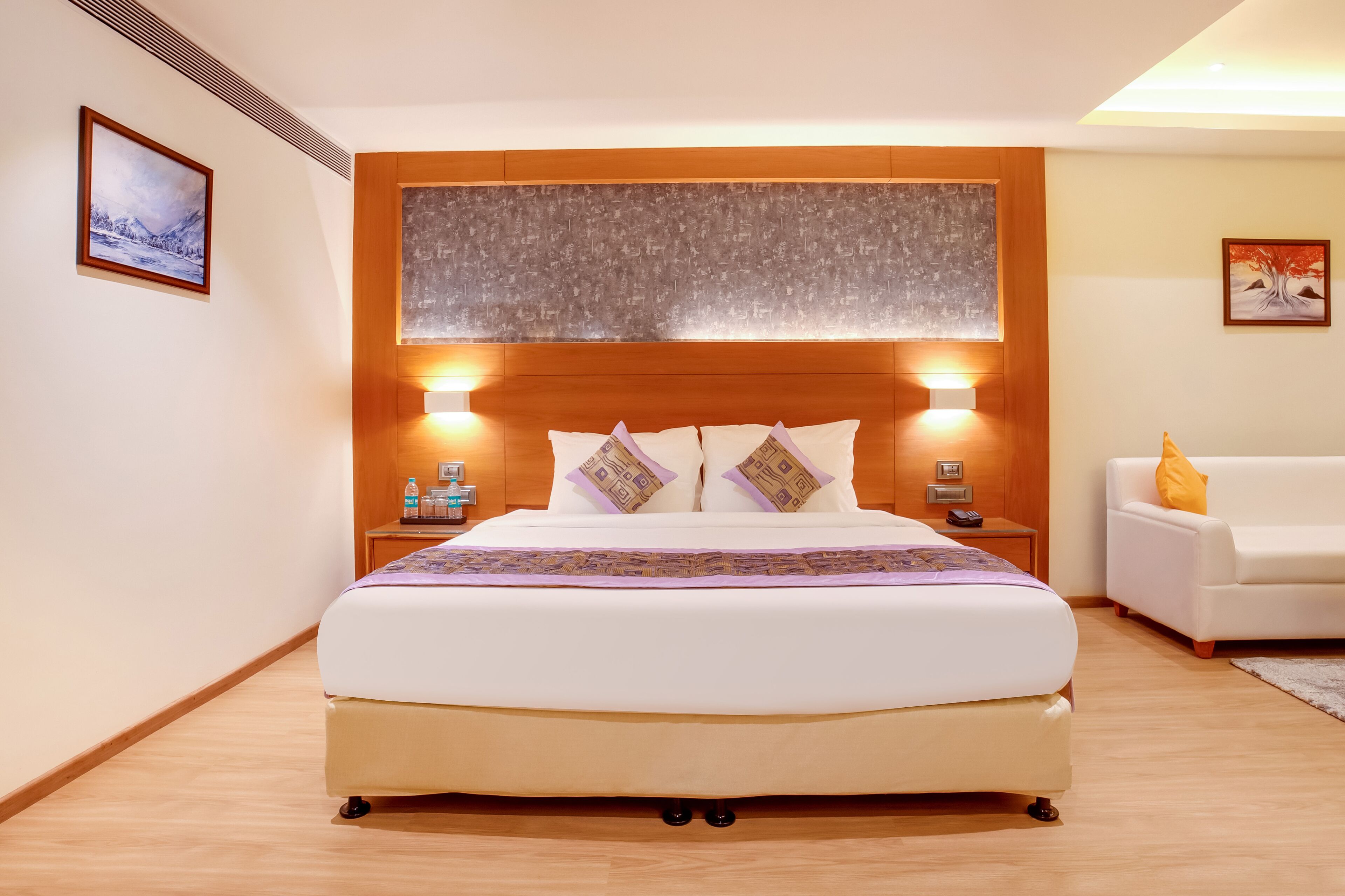 Une chambre d’hôtel avec un grand lit, une tête de lit en bois, des tables de chevet avec des lampes, un canapé blanc et des œuvres d’art accrochées aux murs.