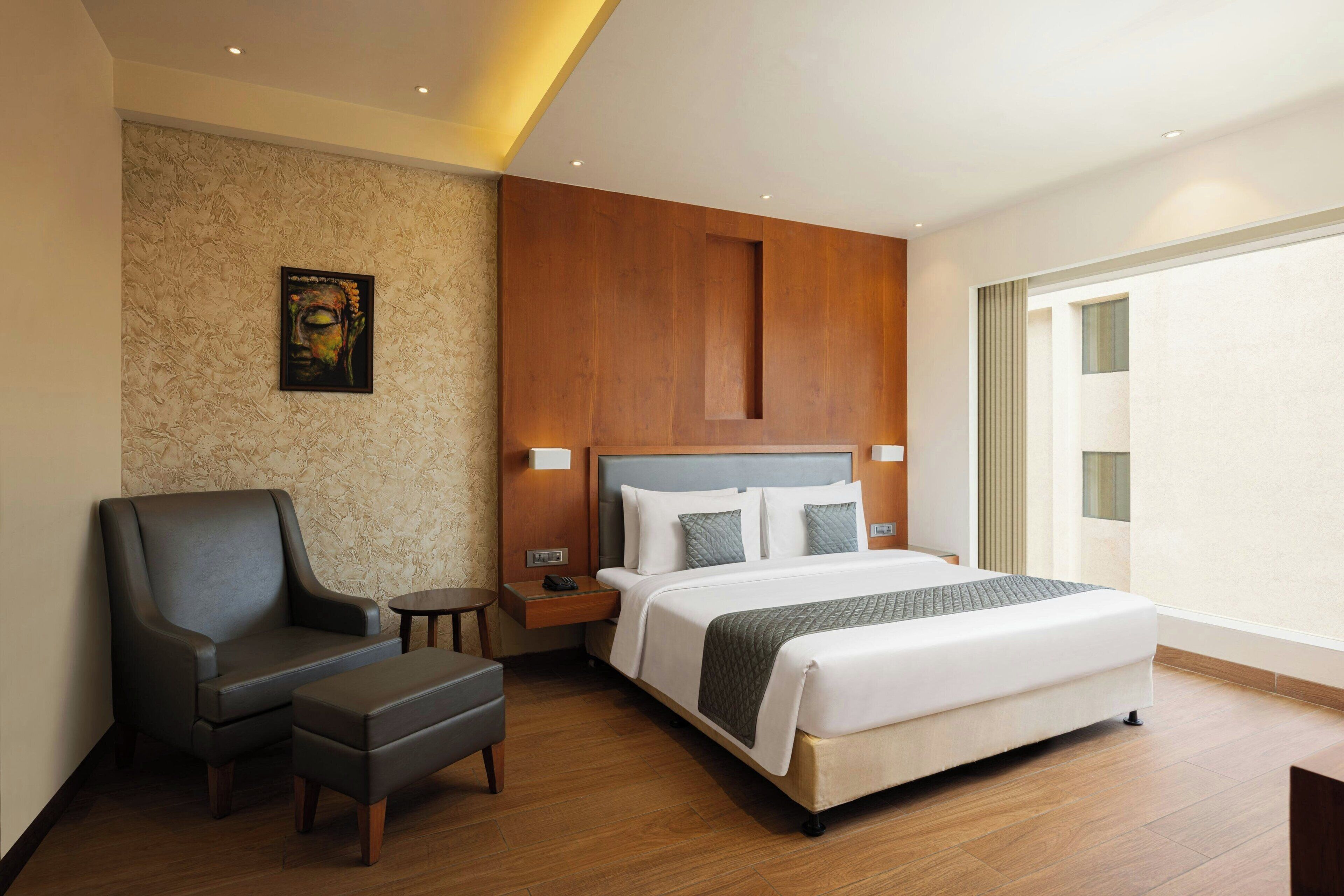 Una habitación de hotel moderna con una cama grande, una zona de estar con un sillón y reposapiés, una mesita y una obra de arte colgada en la pared.