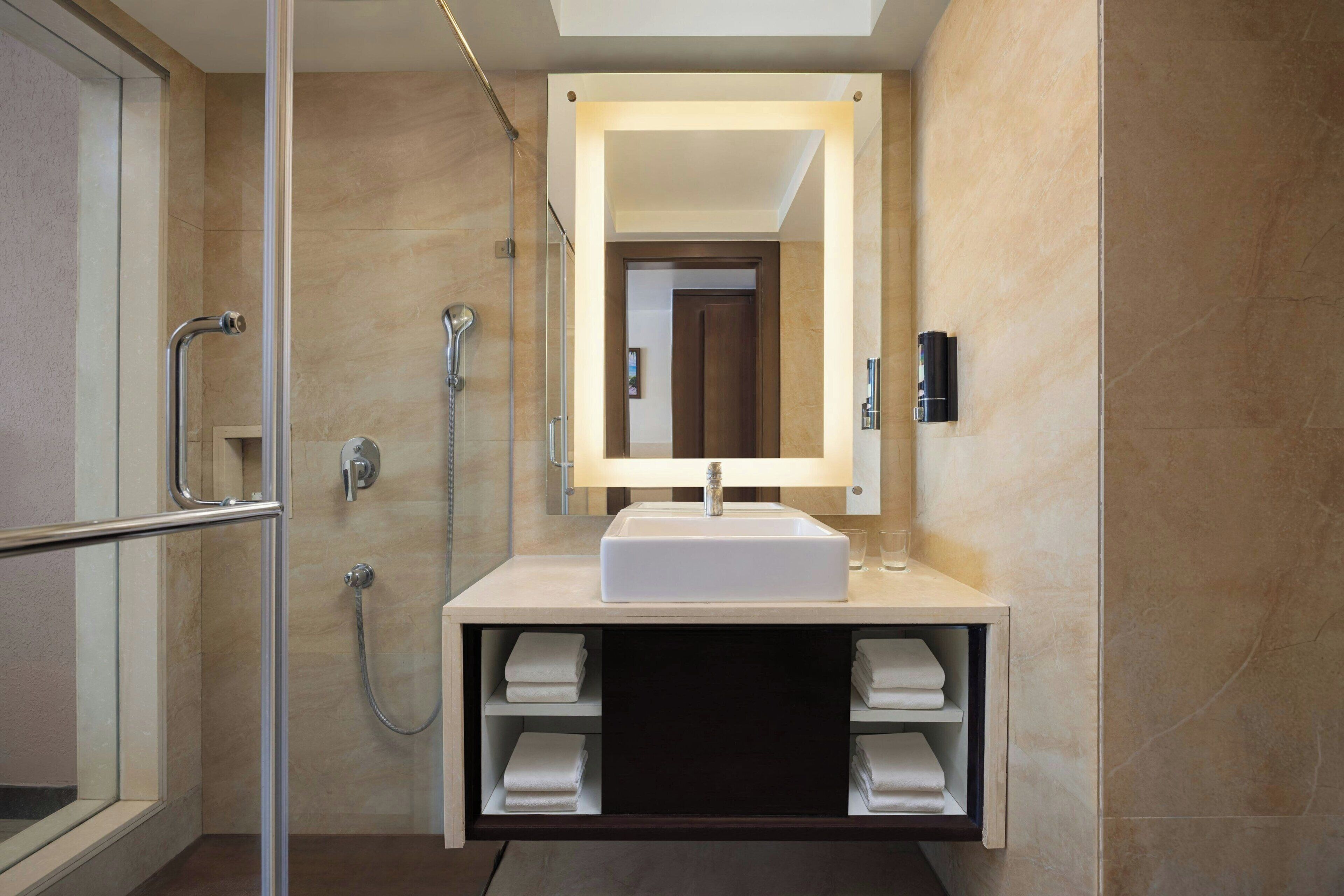 A modern bathroom with a glass-enclosed shower, a white sink, and a mirror above it. There are towels stored in a cabinet below the sink.