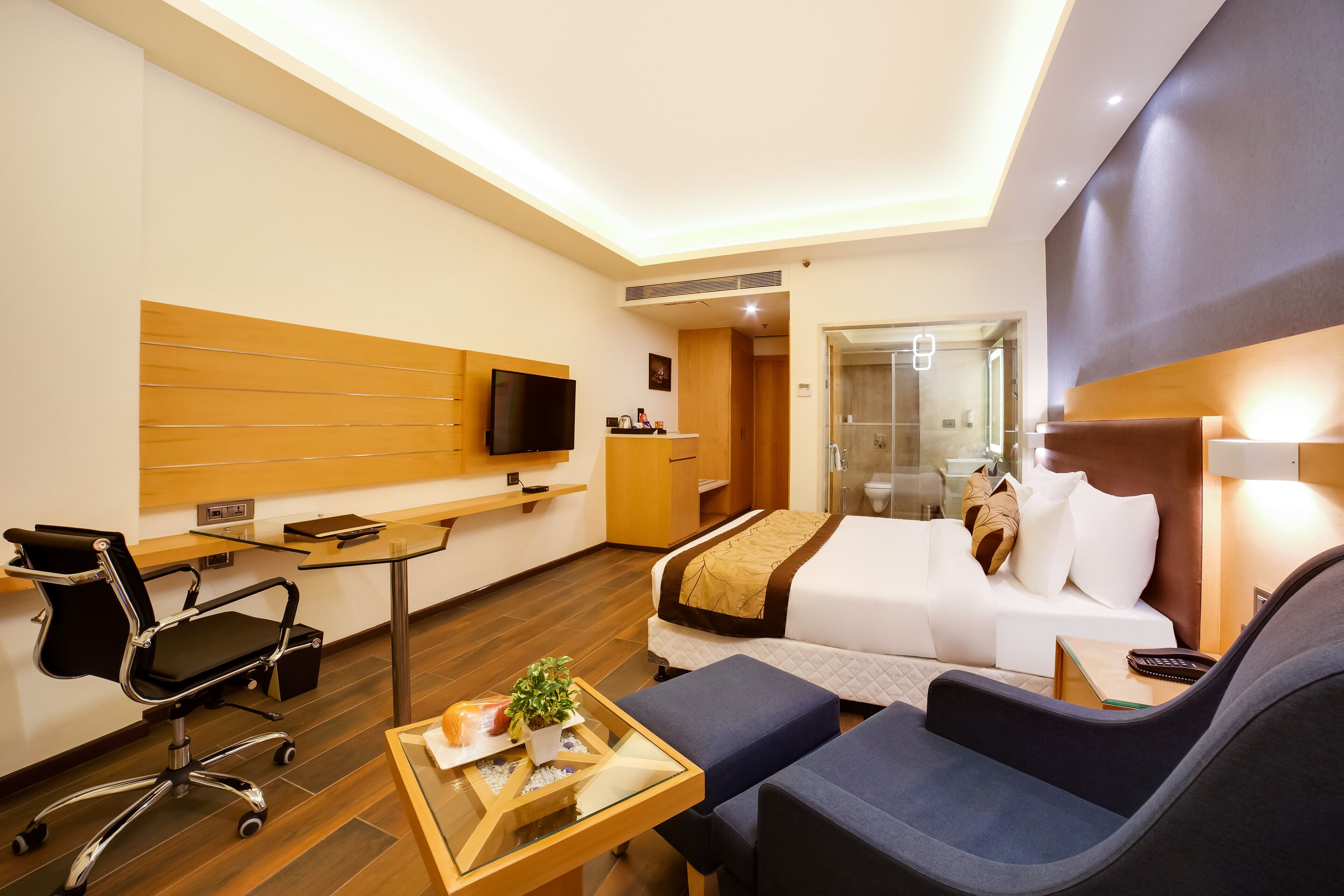 Una habitación de hotel moderna con cama, sofá, escritorio, silla, televisión y zona de ducha de vidrio.
