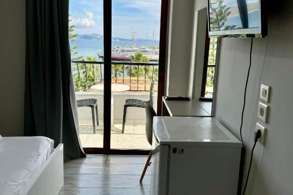 A hotel room with a bed, a desk, a television, and a view of the sea and boats through a sliding glass door.
