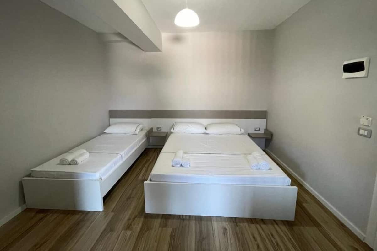 A hotel room with two single beds, a wooden floor, a ceiling light, and a small wall-mounted air conditioning unit.