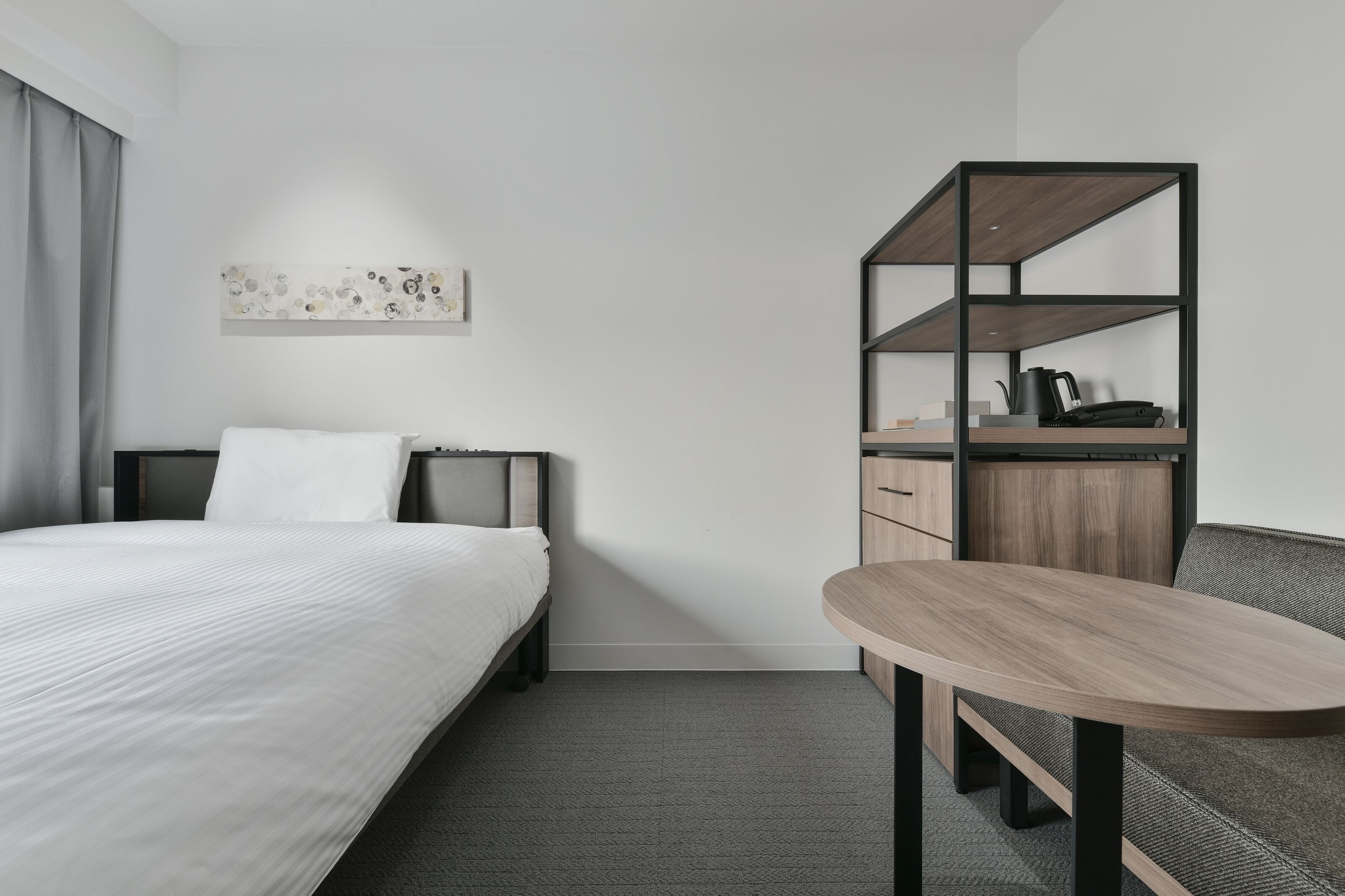 A modern hotel room with a bed, a desk with a chair, and a small kitchenette.