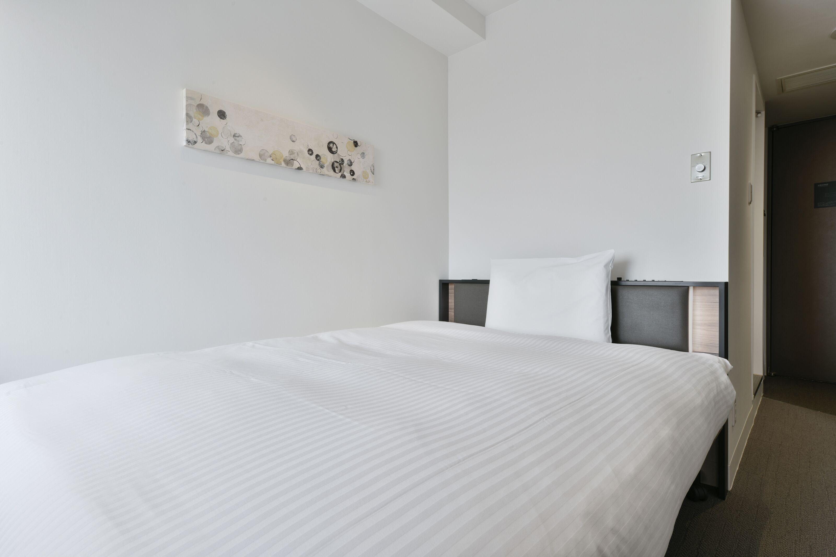 A hotel room with a large bed, a headboard, a wall-mounted artwork, and a door leading to another room.