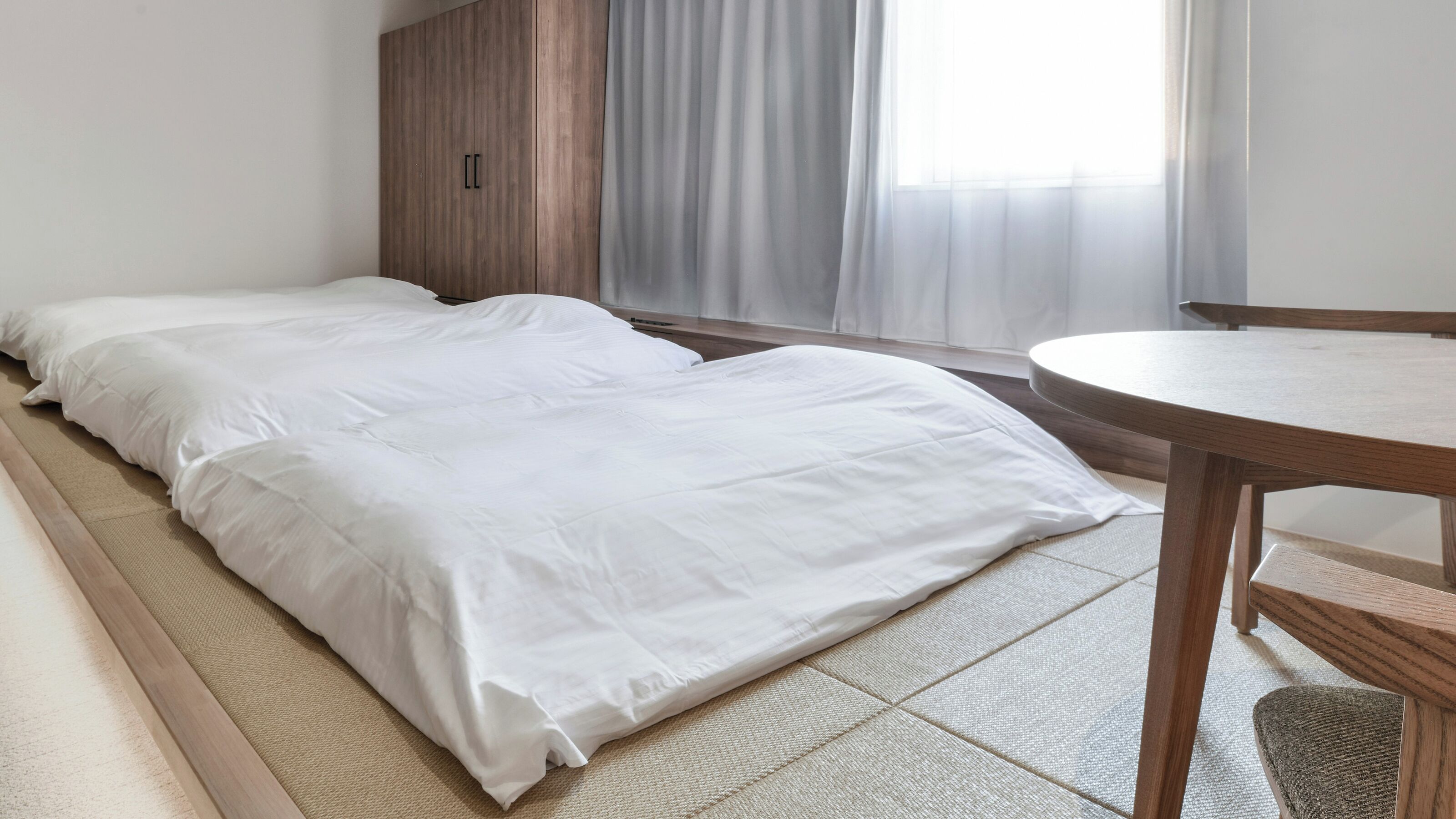 A room with a bed, a wooden wardrobe, a round table, and a window with curtains.