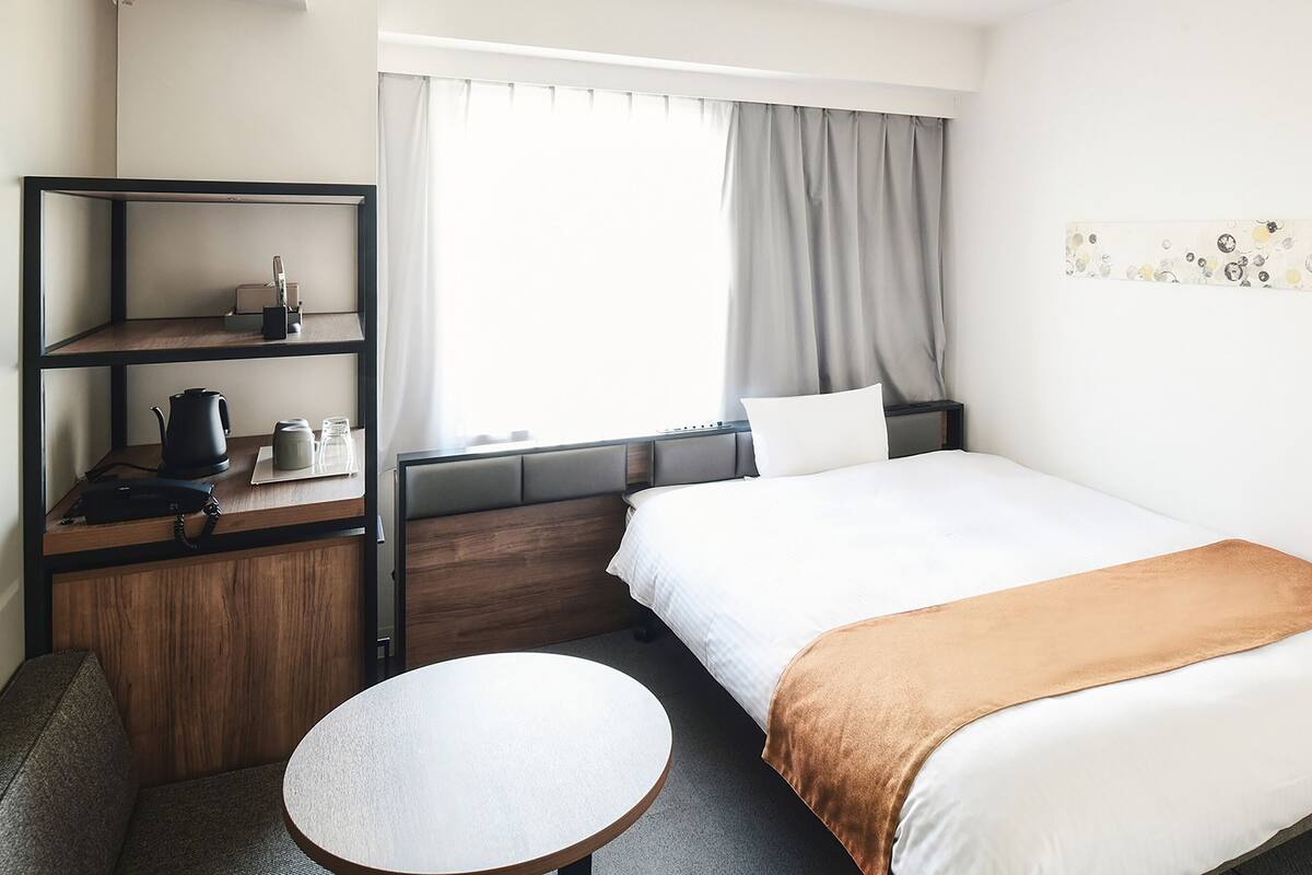 Een hotelkamer met een bed, een klein rond tafeltje, een bureau met een waterkoker, en een plank met een waterkoker en glazen.