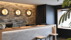 Un espace d’accueil moderne avec un mur en briques, trois appliques murales, un panneau vertical foncé et une fenêtre agrémentée d’une plante.