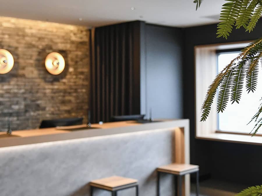 A modern reception area with a brick wall, three wall-mounted lights, a dark vertical panel, and a window with a plant.