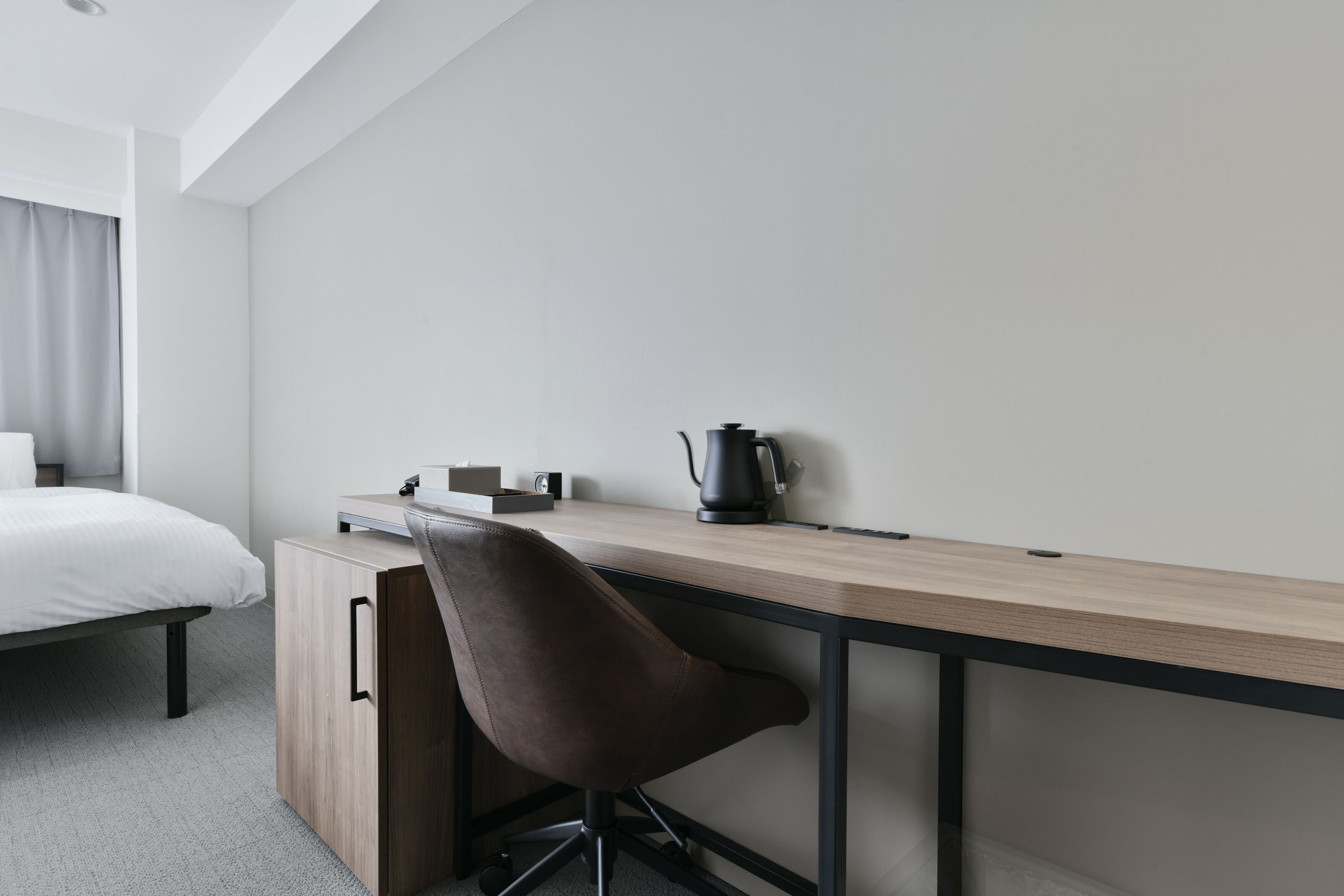 A modern hotel room with a wooden desk, a brown chair, a bed with white bedding, and a kettle on the desk.