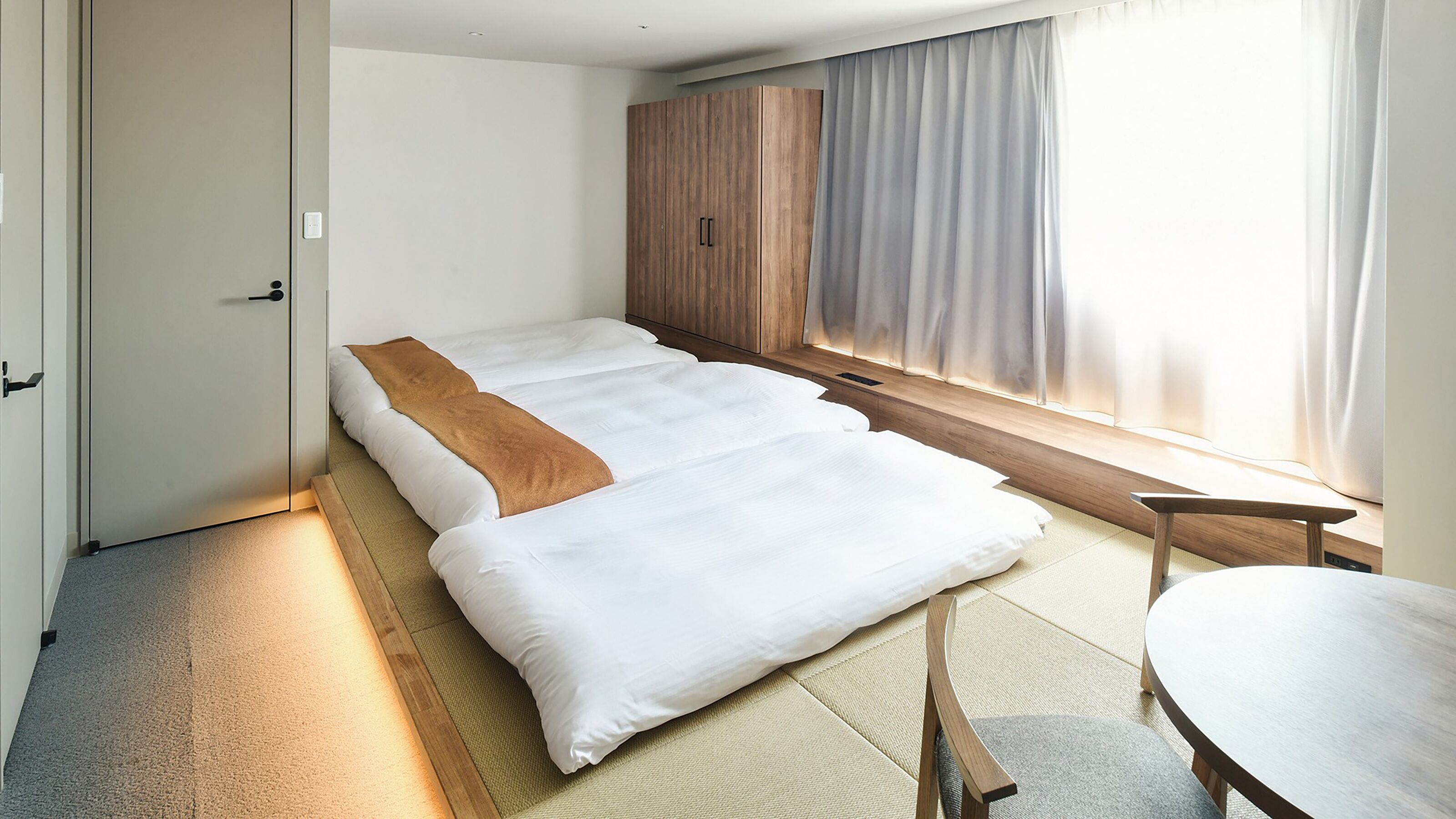 A minimalist hotel room with a large bed, a wooden wardrobe, a small table, and a window with curtains.