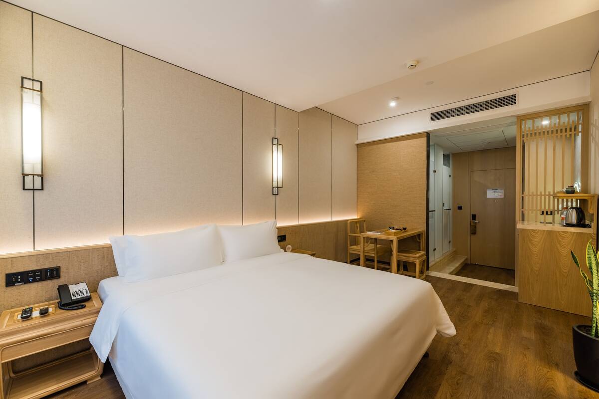 A modern hotel room with a large bed, bedside table with a phone, and a desk with a chair.