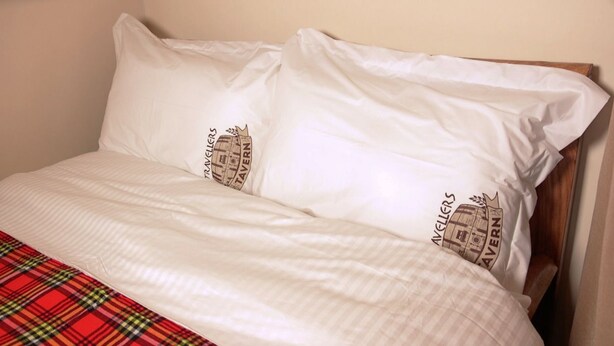 A bed with white bedding and two pillows featuring a logo. - Travellers Tavern (Nkubu)
