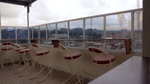 A balcony with four red cushioned chairs and a table, overlooking a cityscape with buildings and a tower. - Travellers Tavern (Nkubu)