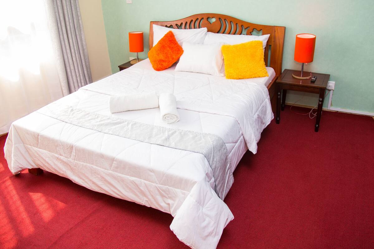 A neatly made bed with white bedding and two pillows, one orange and one yellow, in a room with red carpet and light green walls.