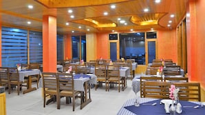 A restaurant with wooden tables and chairs, arranged in a spacious layout with large windows and a high ceiling. - Gezellig Inn - Kanyal Villas (Manali)