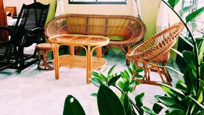 Une pièce comprenant un ensemble de meubles de salon en osier, une petite table ronde et une chaise noire. On y trouve également des plantes vertes et une fenêtre avec des stores.