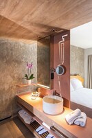 A modern bathroom with a wooden vanity, a round sink, a mirror, a potted orchid, and a bed with white linens.