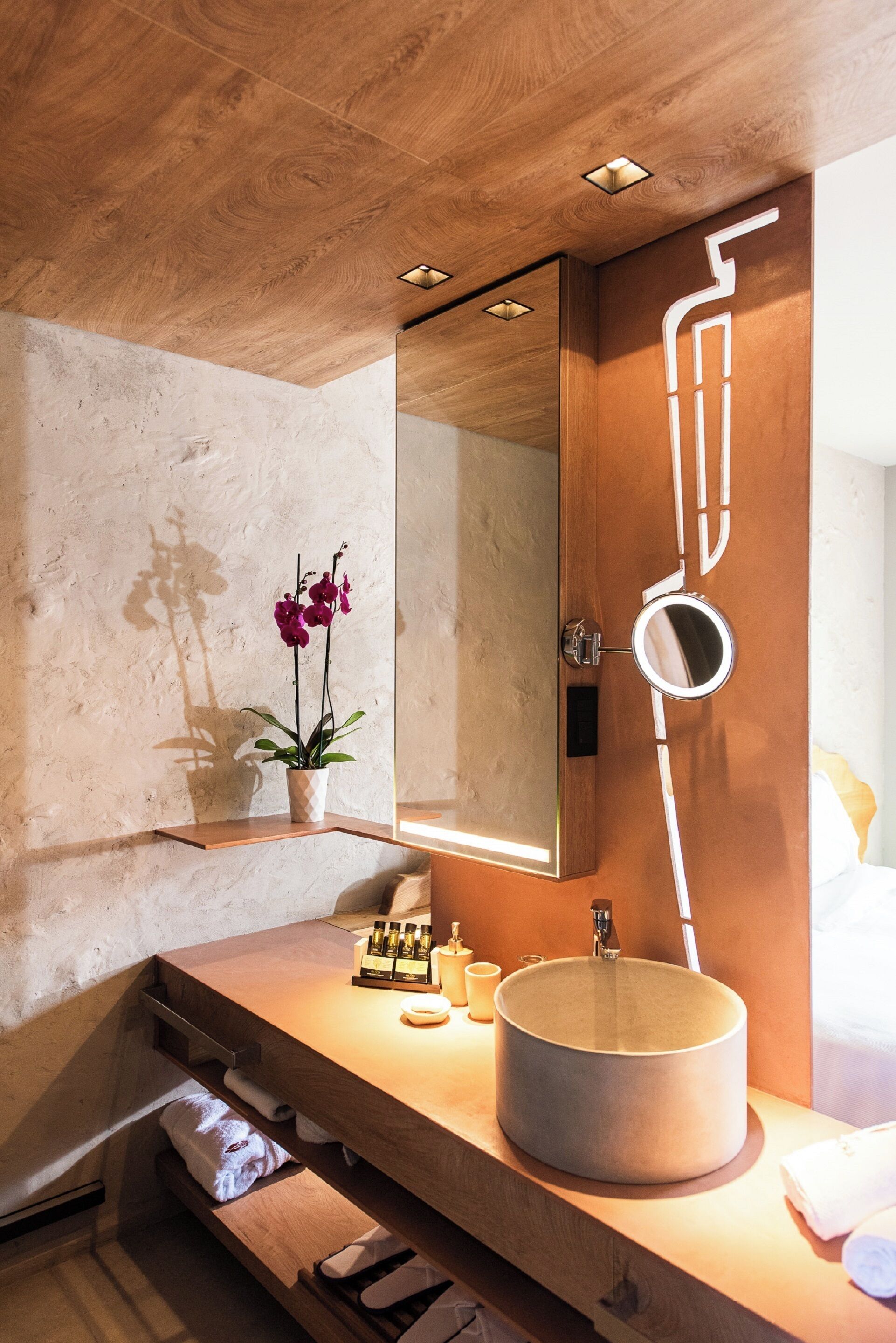 Ein modernes Badezimmer mit einem Holzkosmetikschrank, einem runden Waschbecken, einem Spiegel mit einem außergewöhnlichen Griff und einer Topf-Orchidee.