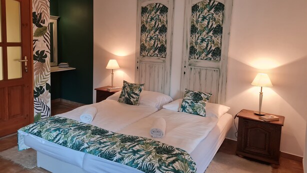 Une chambre à coucher comprenant un lit, deux tables de chevet, une porte et un mur orné d’un motif de feuilles tropicales.