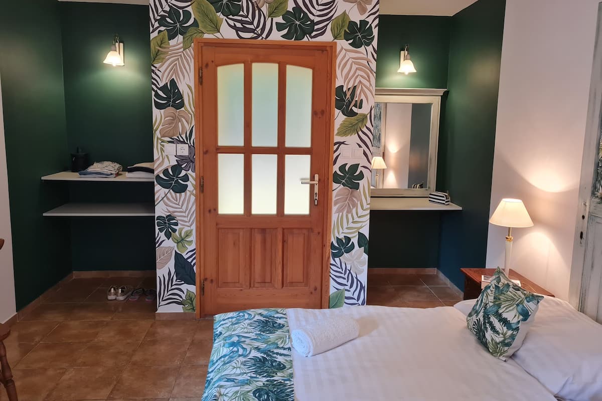 A bedroom with a wooden door, a bed with a tropical print bedspread, a nightstand with a lamp, and a wall-mounted shelf with a mirror.