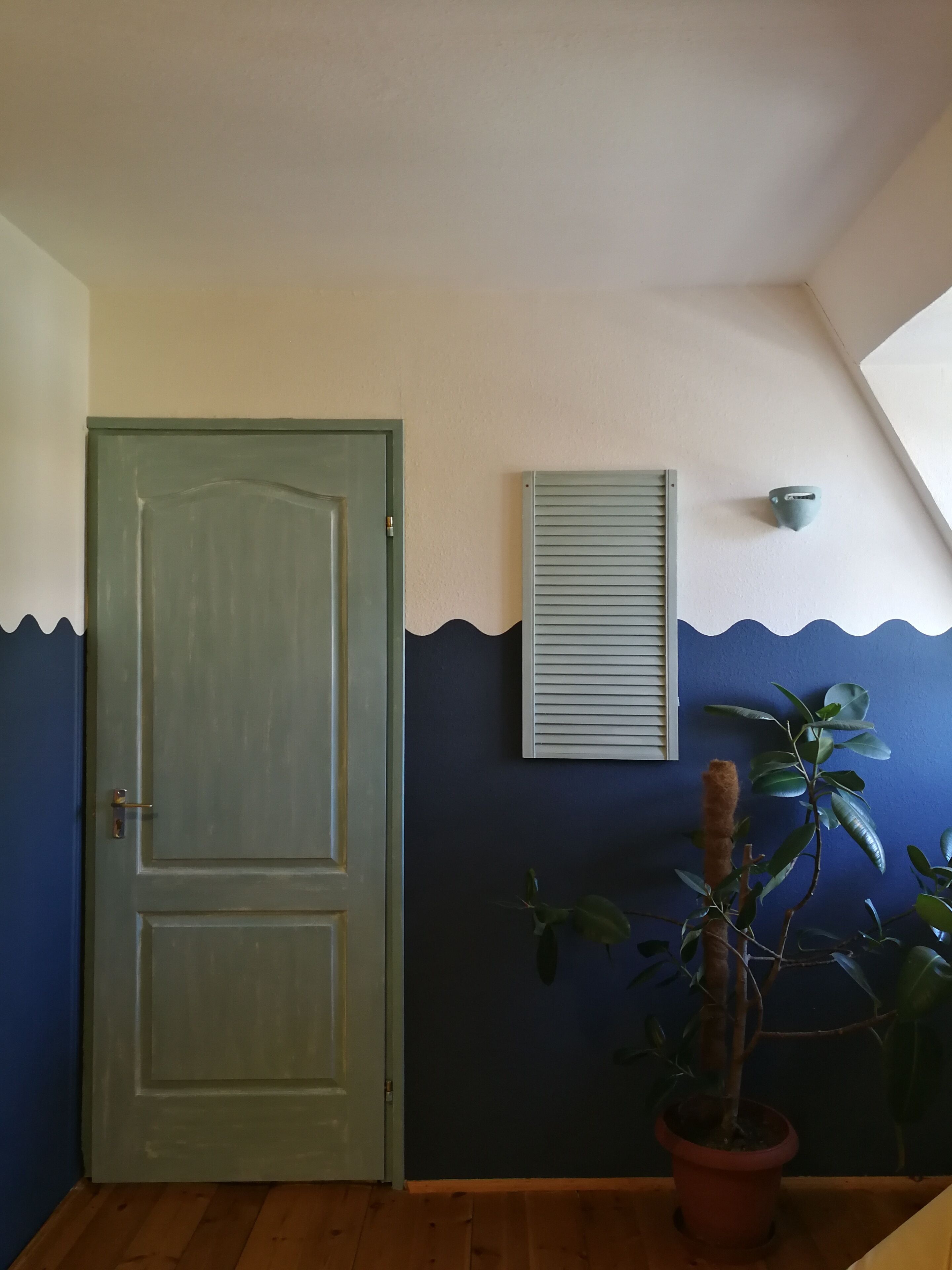 A room with a wooden floor, a green door, a blue wall with a wave pattern, a potted plant, and a wall-mounted air conditioning unit.