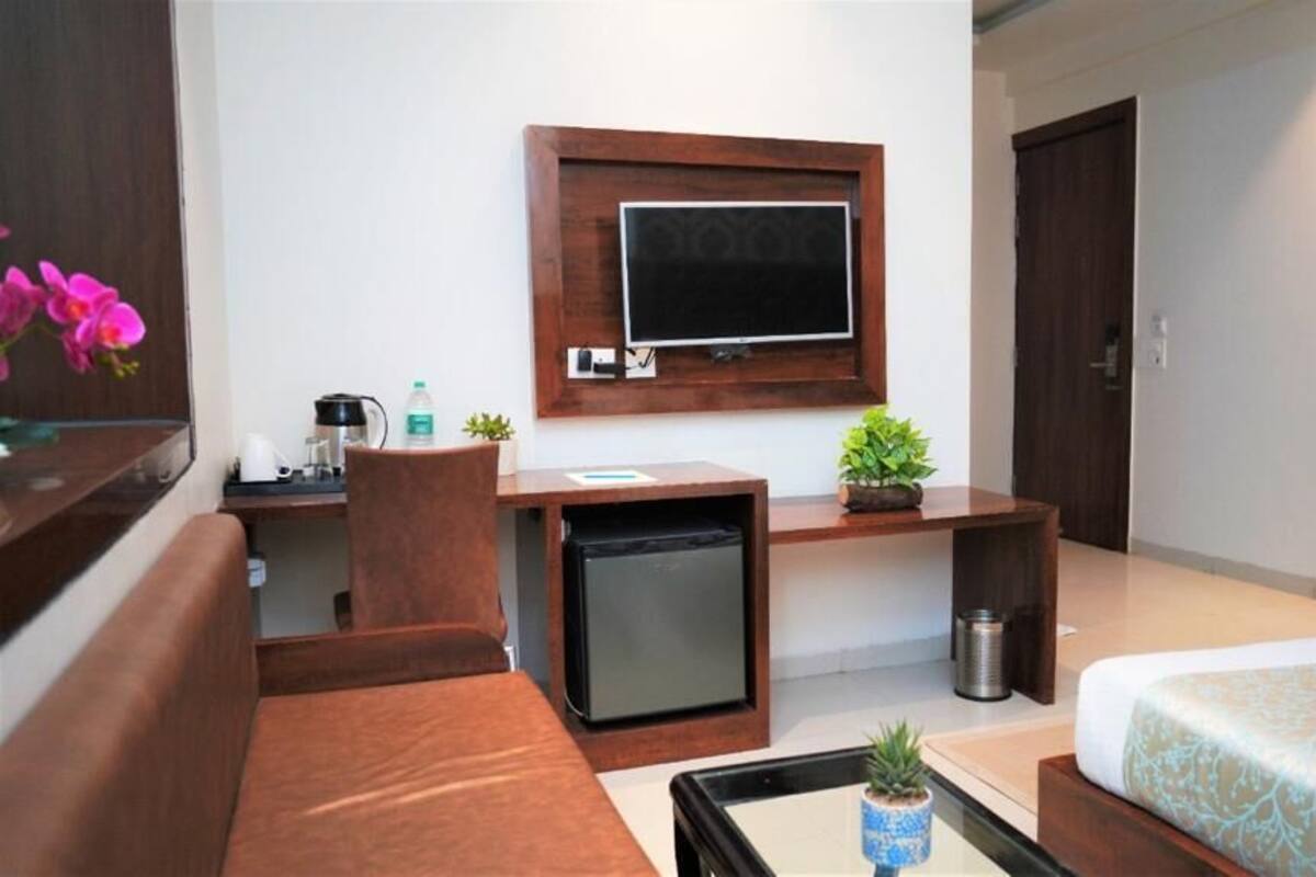 A hotel room with a bed, a desk with a television, a small refrigerator, a sink, and a potted plant.