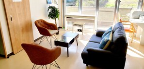 A modern living room with a sofa, armchair, and coffee table. There is a potted plant and a view of the outdoors through large windows.