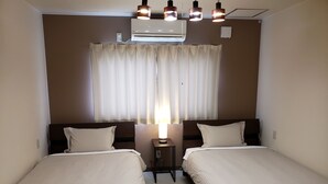 A hotel room with two single beds, a bedside table with a lamp, an air conditioning unit, and a window with curtains. - Resort Inn Gabusoka (Nago)