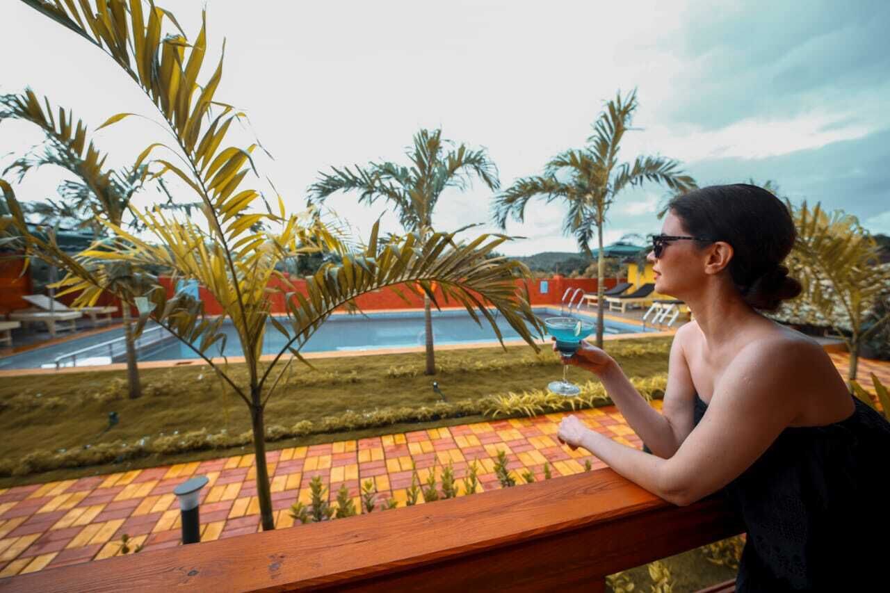 Eine Person sitzt auf einer Holzterrasse mit Blick auf ein Schwimmbecken, vorbei an Palmen und einem roten Gebäude im Hintergrund.