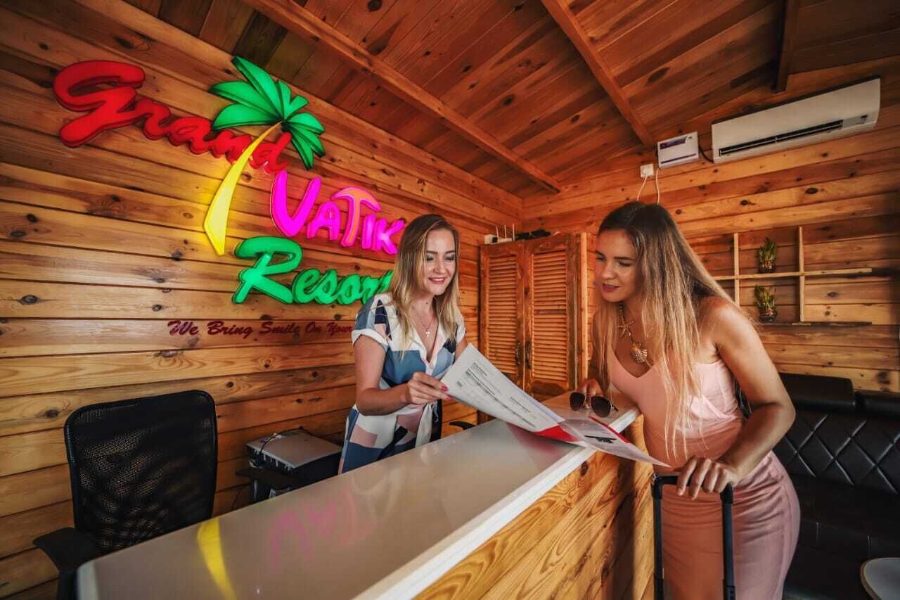 A wooden interior with a reception desk, a neon sign reading "Grand Vatik Resort," and a black leather couch.